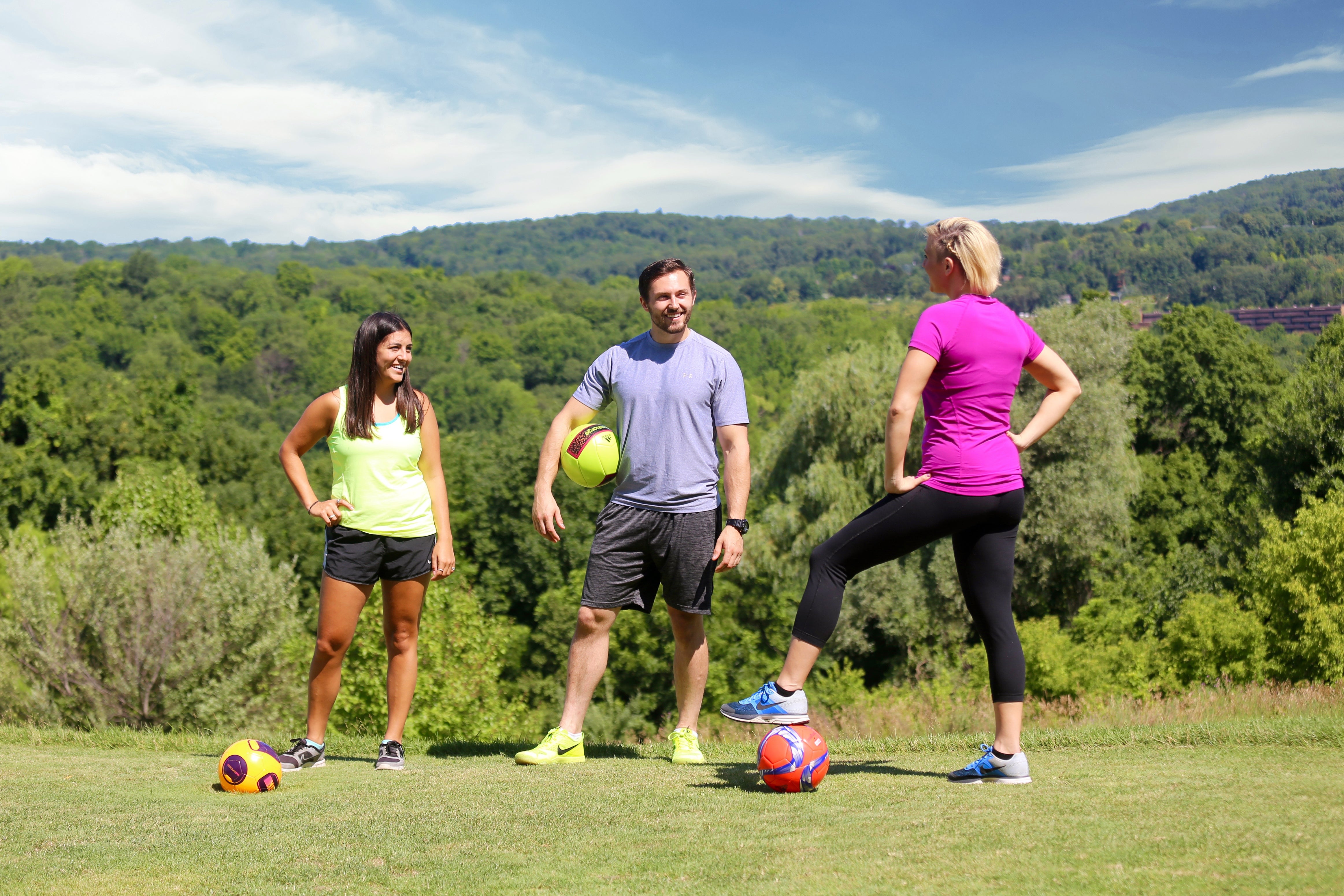 Three people standing on a golf course with soccer balls ready to play foot golf