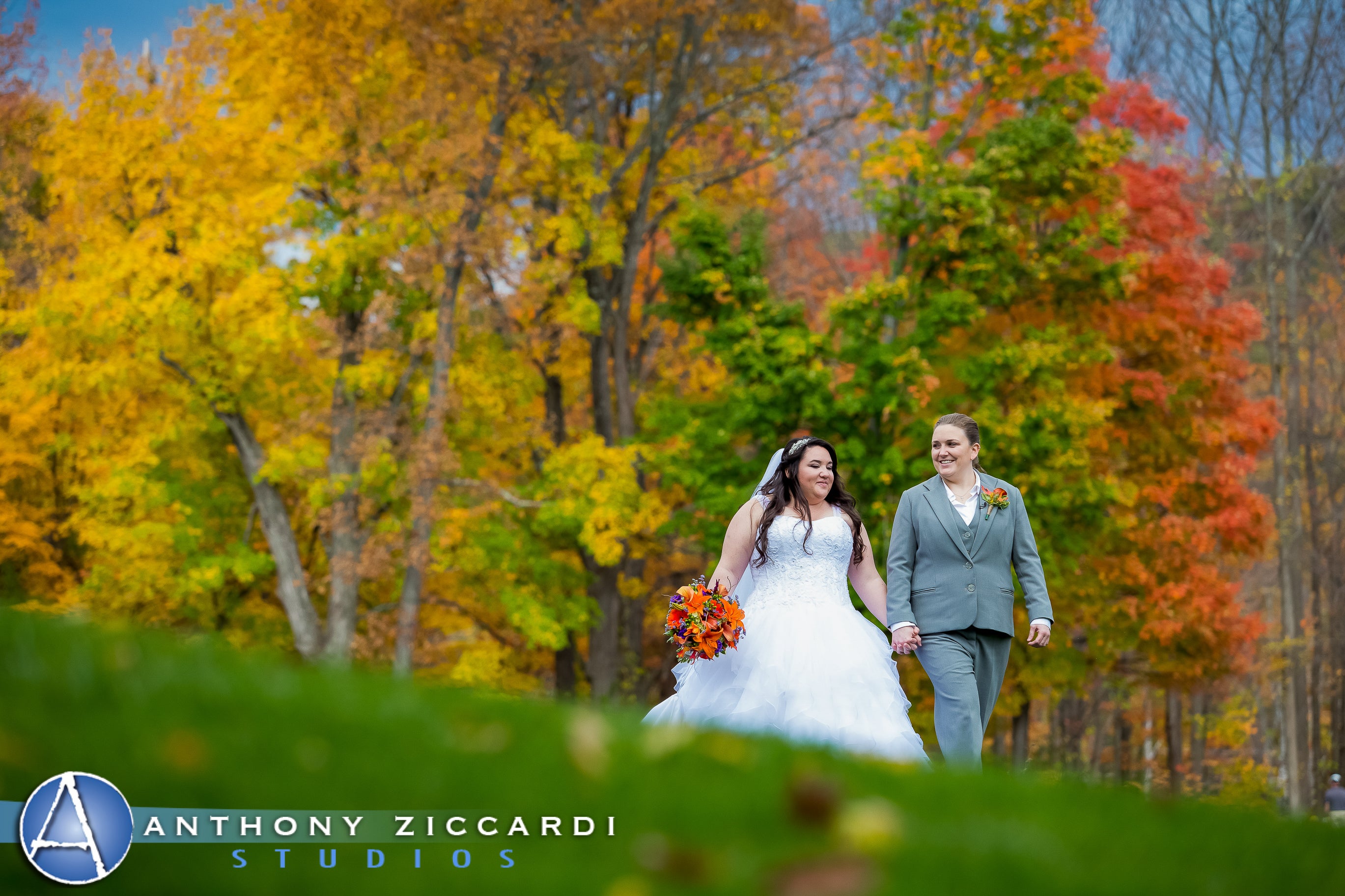 Bride and bride walking hand in hand with fall trees behind them.