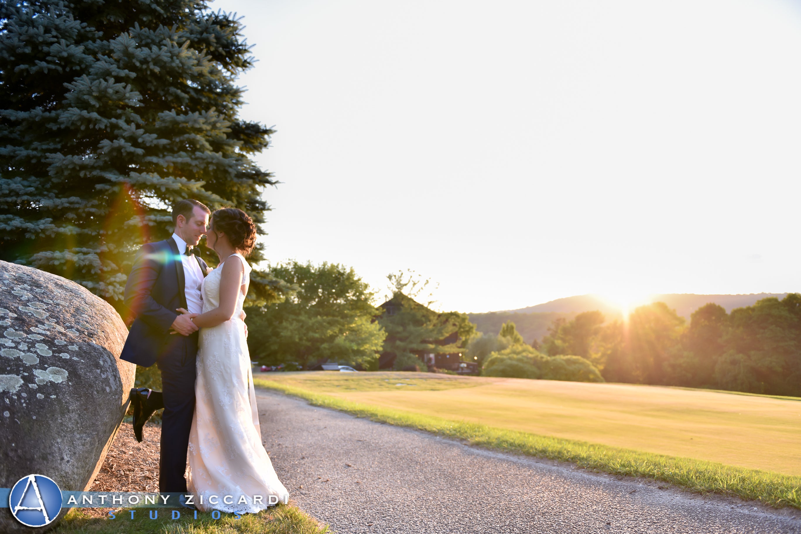 Bride and groom lean against a large rock as sun sets behind them.