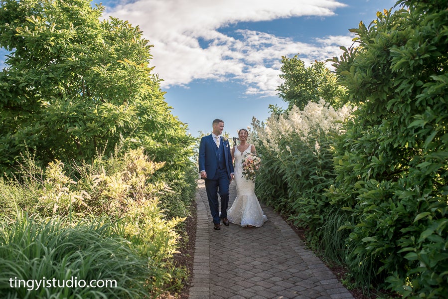 Bride and groom walk down pathway surrounded by greenery.
