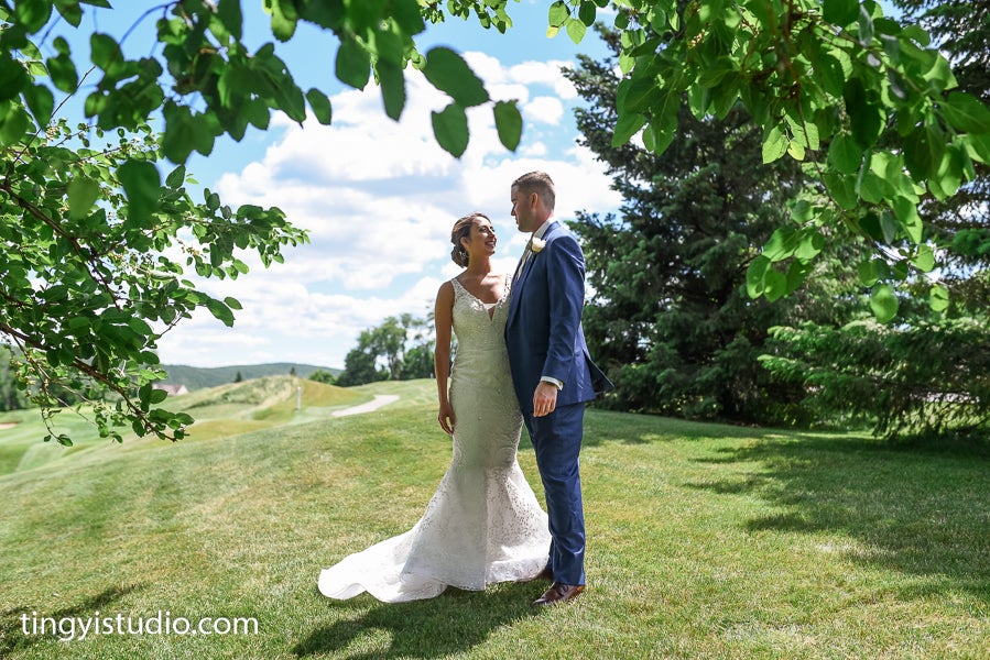Bride and groom stand side by side between trees.