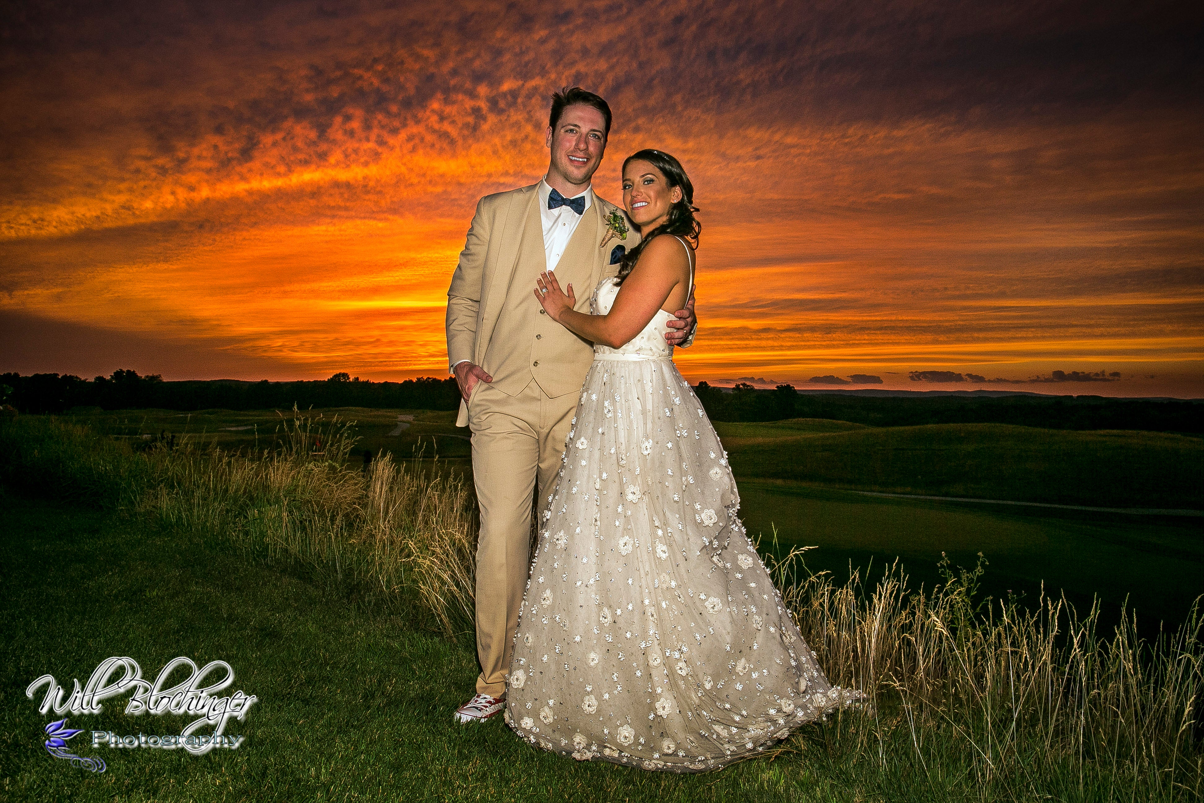 Bride and groom, who is wearing a tan tux, stand in front of orange sunset.
