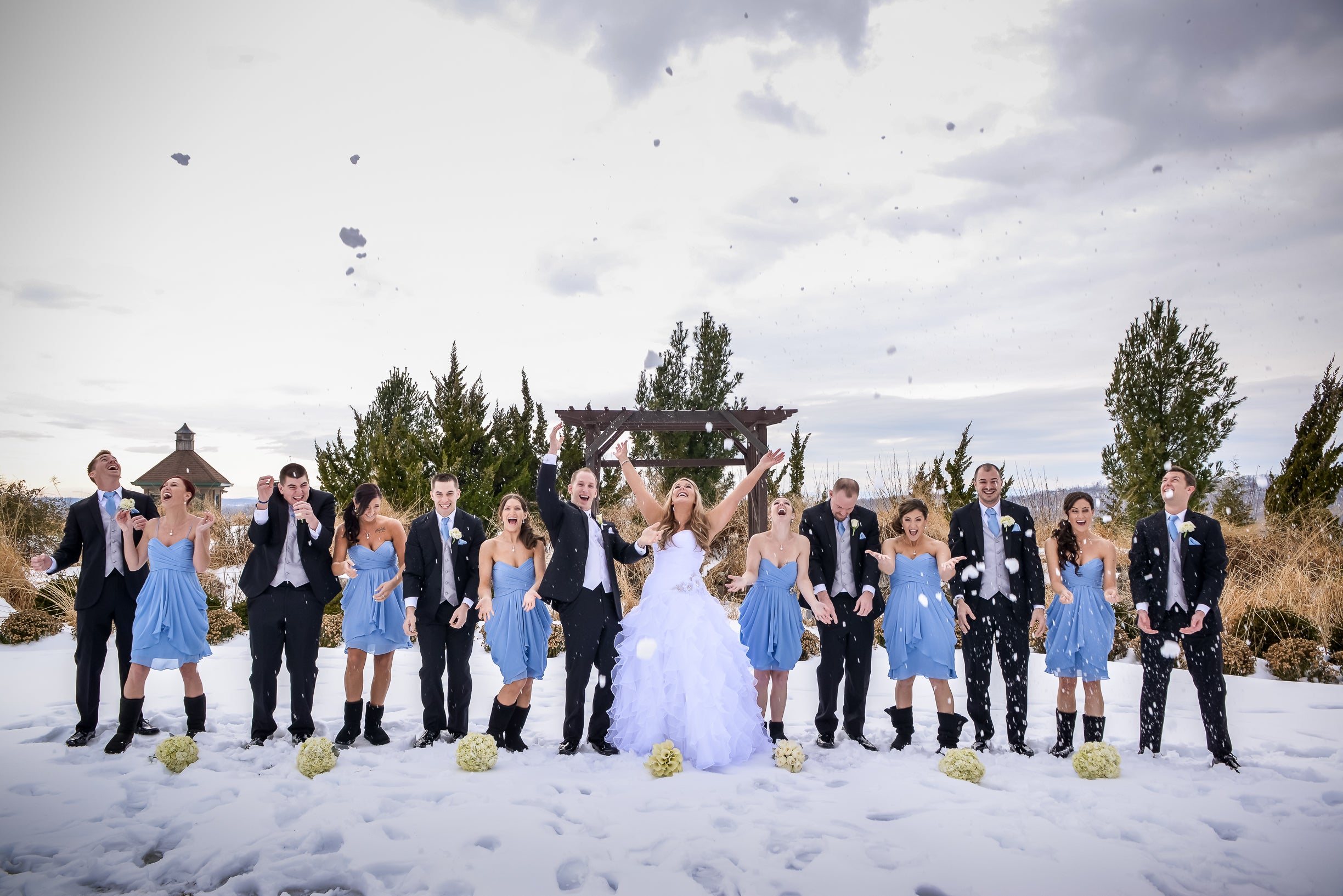 Wedding party, featuring men in black tuxes and bridesmaids in short blue dresses, stand together in a line and throw snow in air.