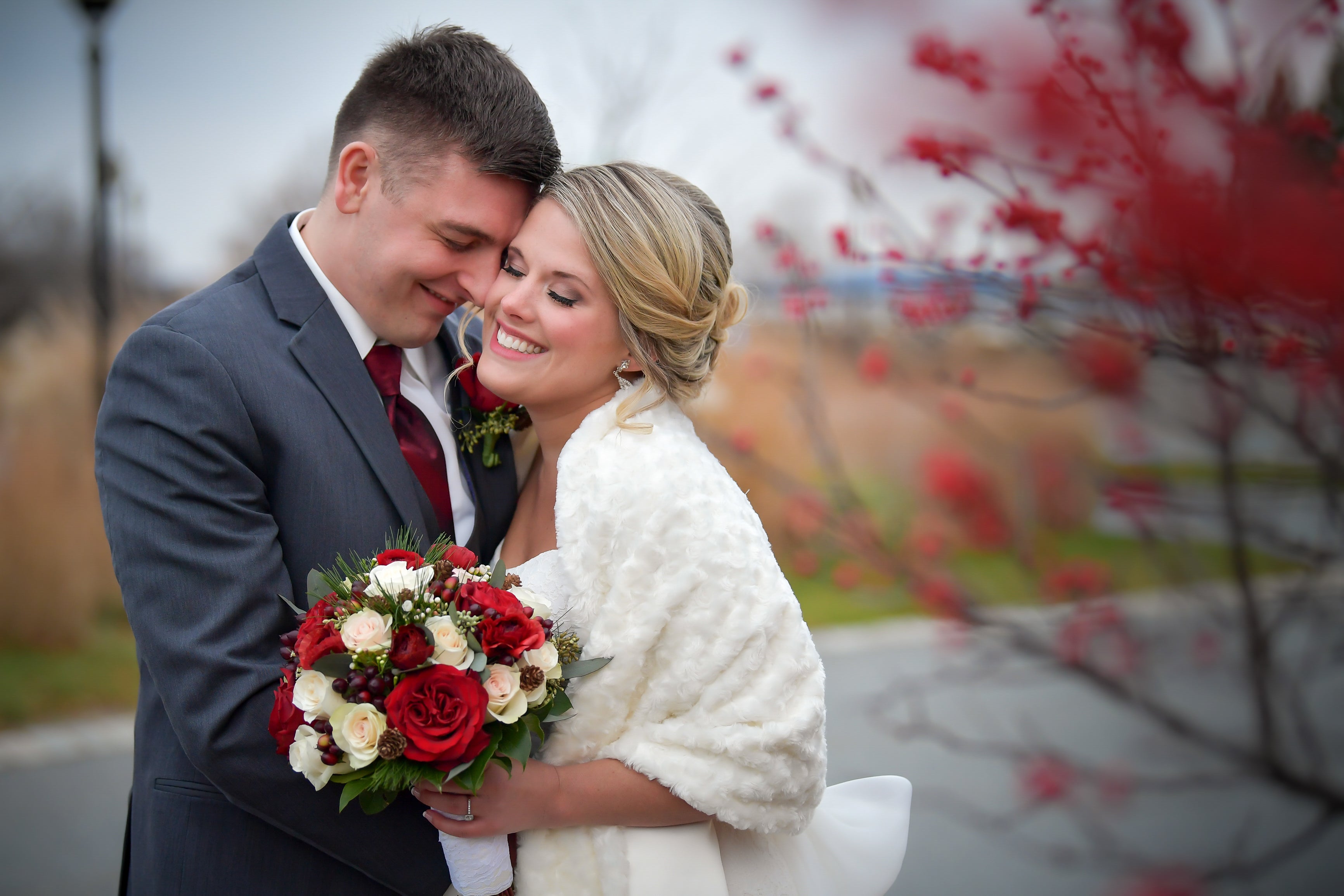 Bride and groom stand together with eye closed as bride holds bouquet of white and red roses.