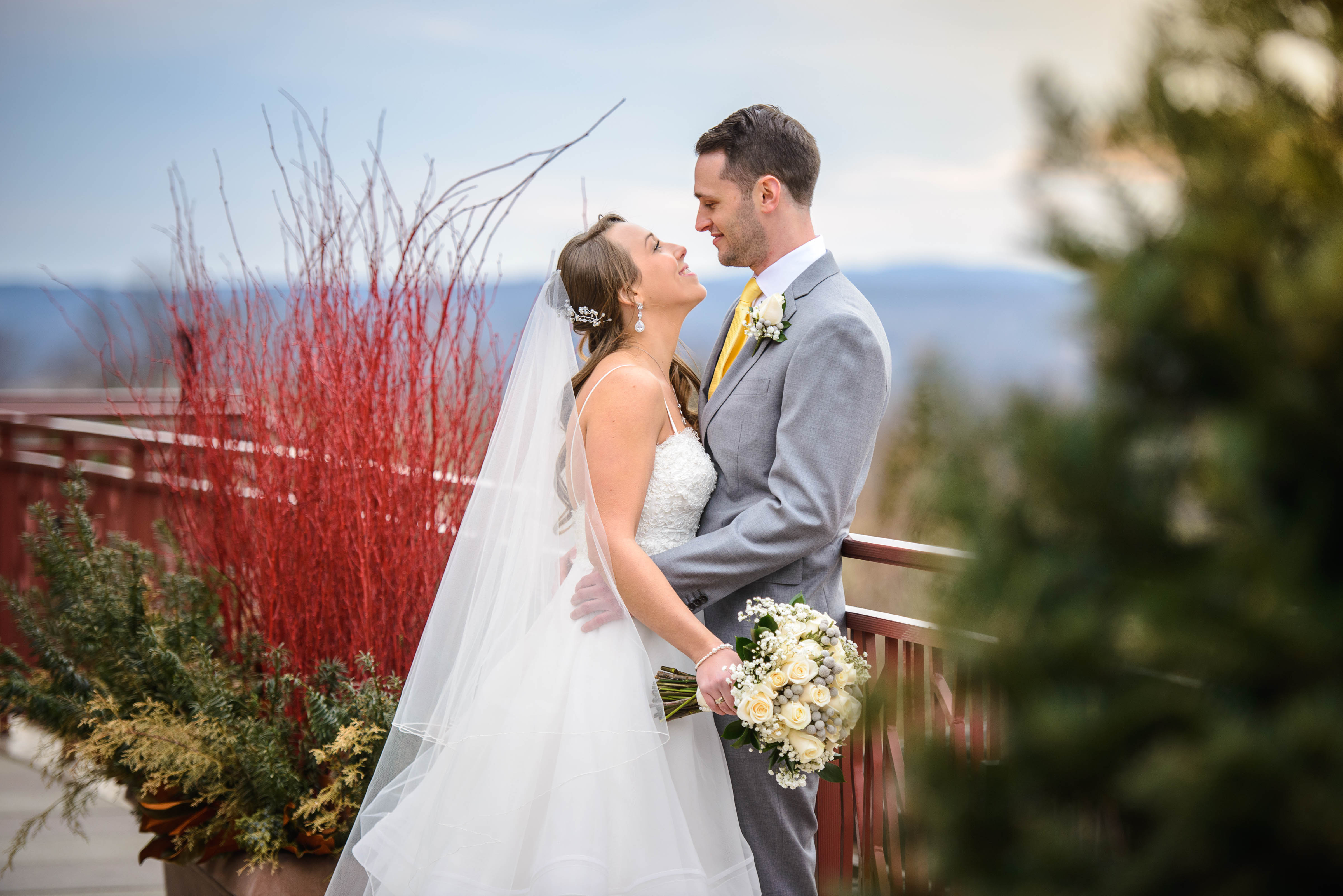 Bride and groom look at one another as they lean on railing with mountains behind them.