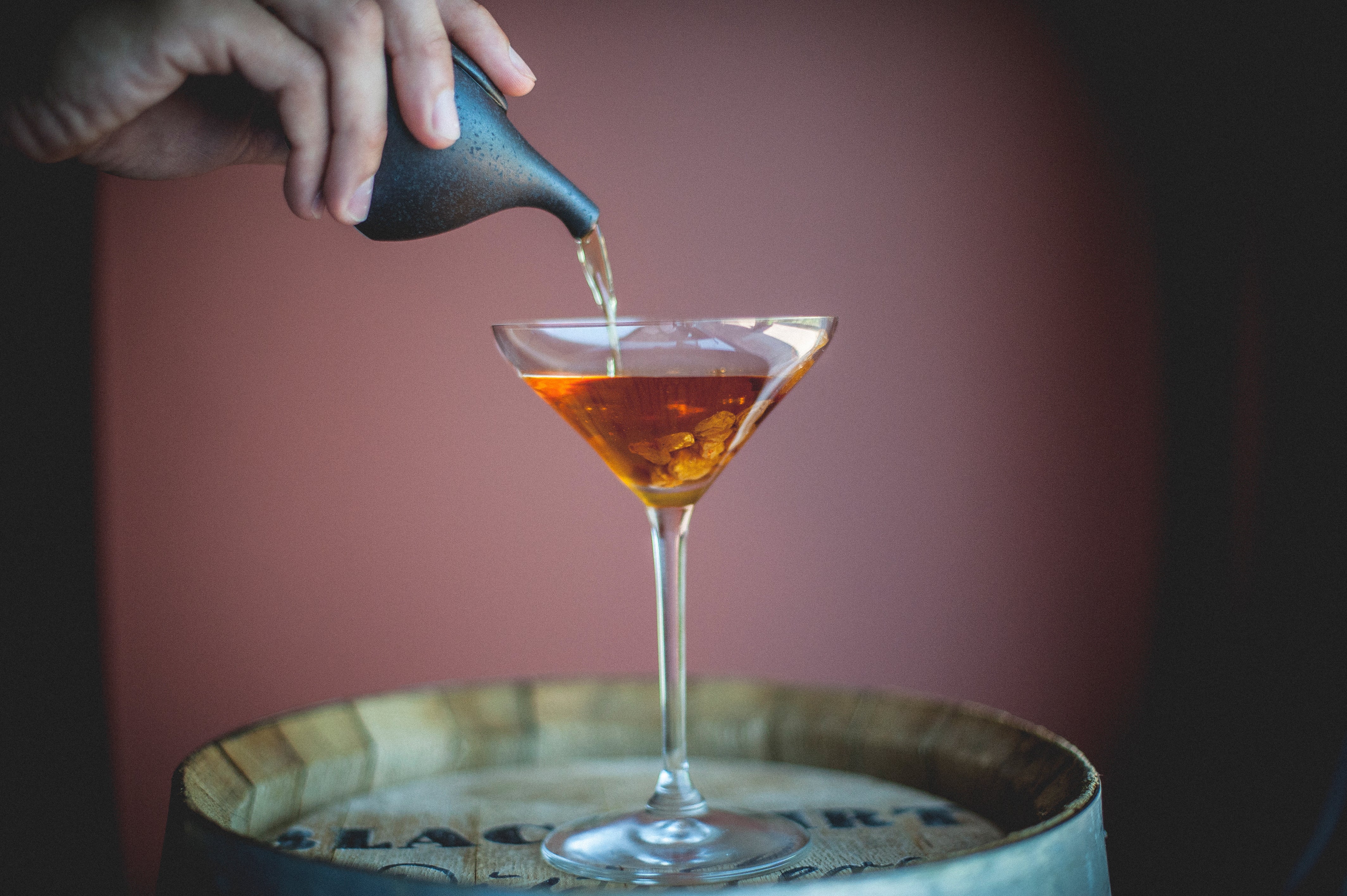 Hand pouring cocktail into martini glass. on top of a barrel.