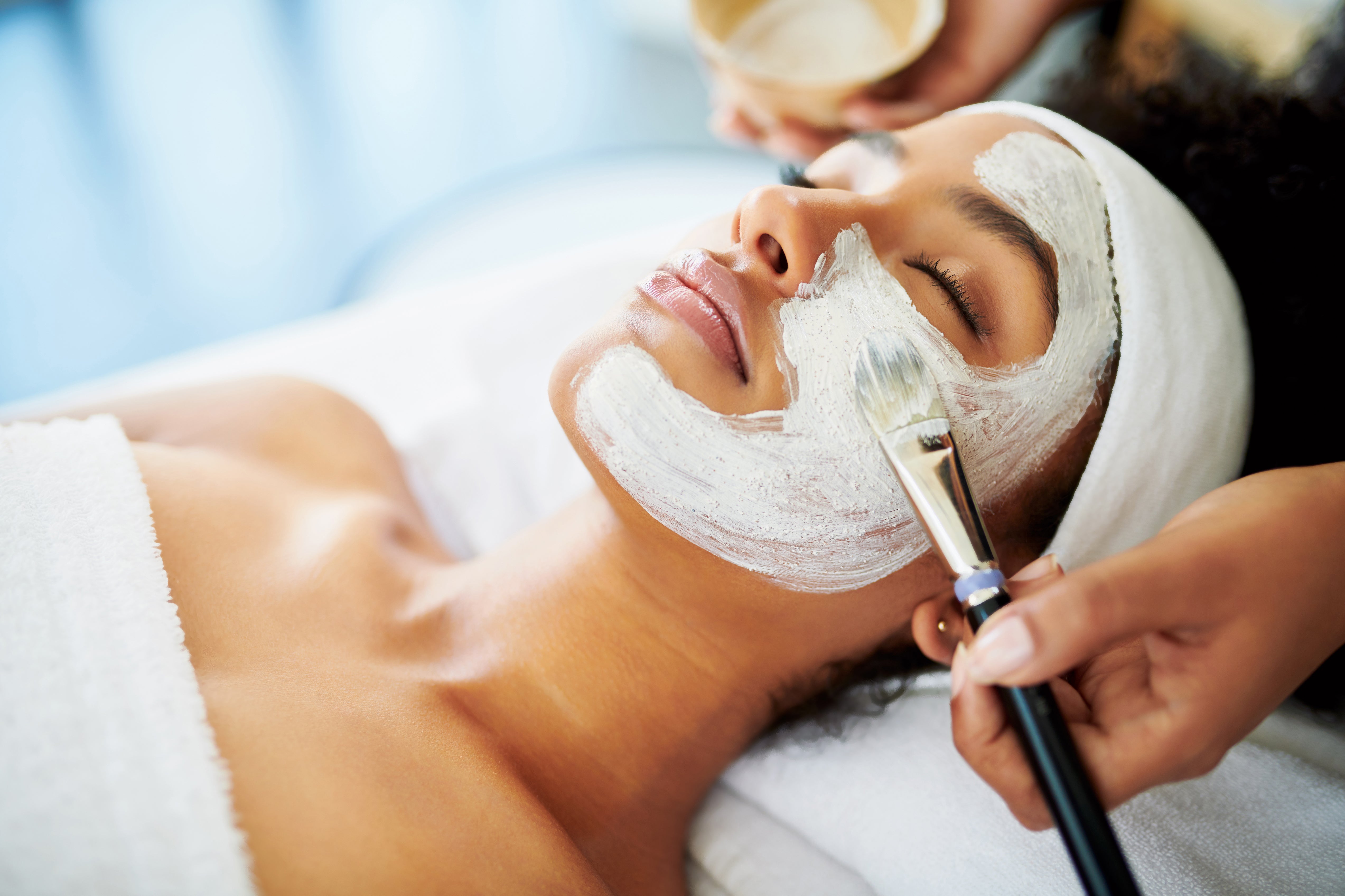 Tech painting white face mask on woman's face during spa treatment.
