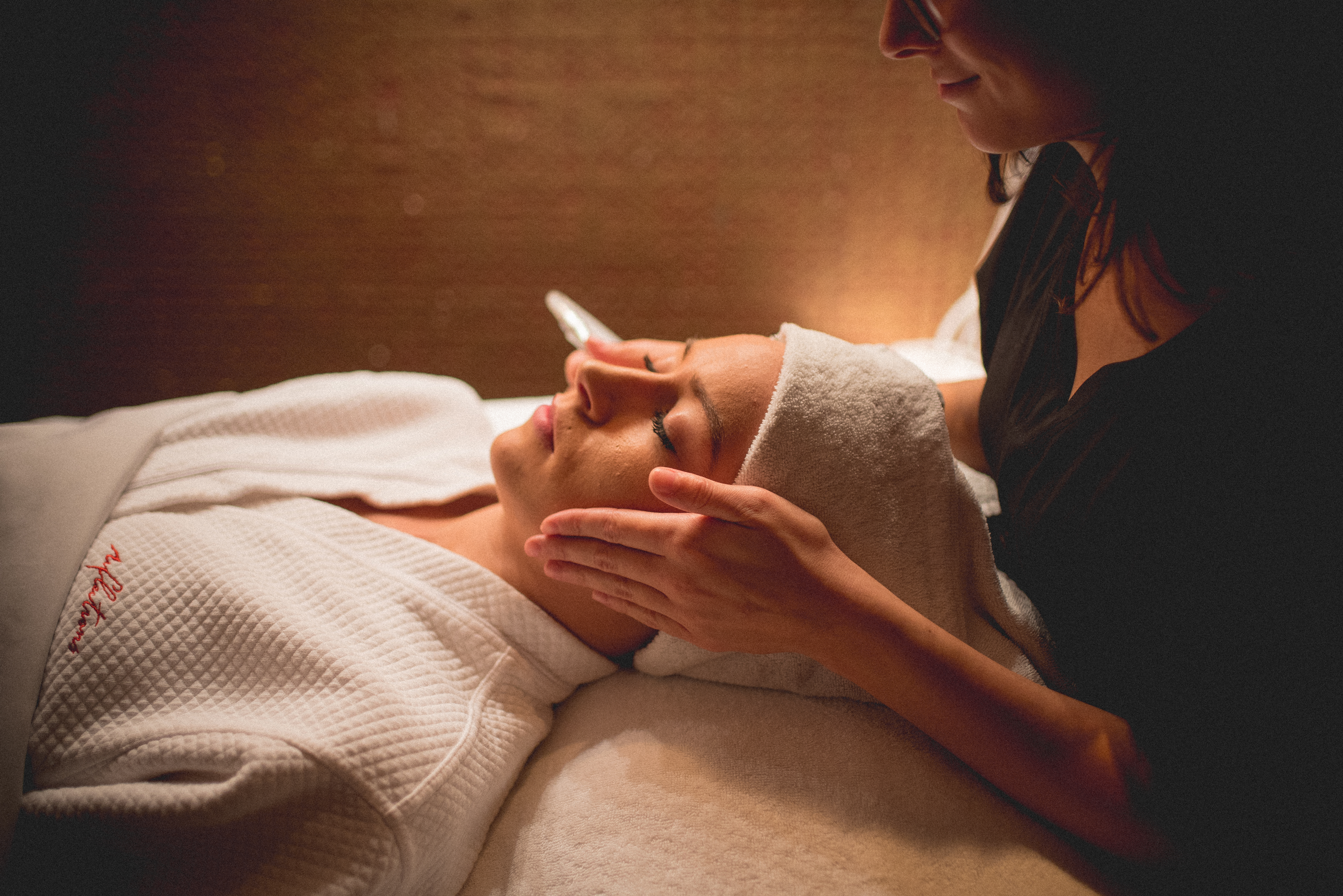 Woman in white robe and white hair towel laying on table with facialists hands on either side of her head.