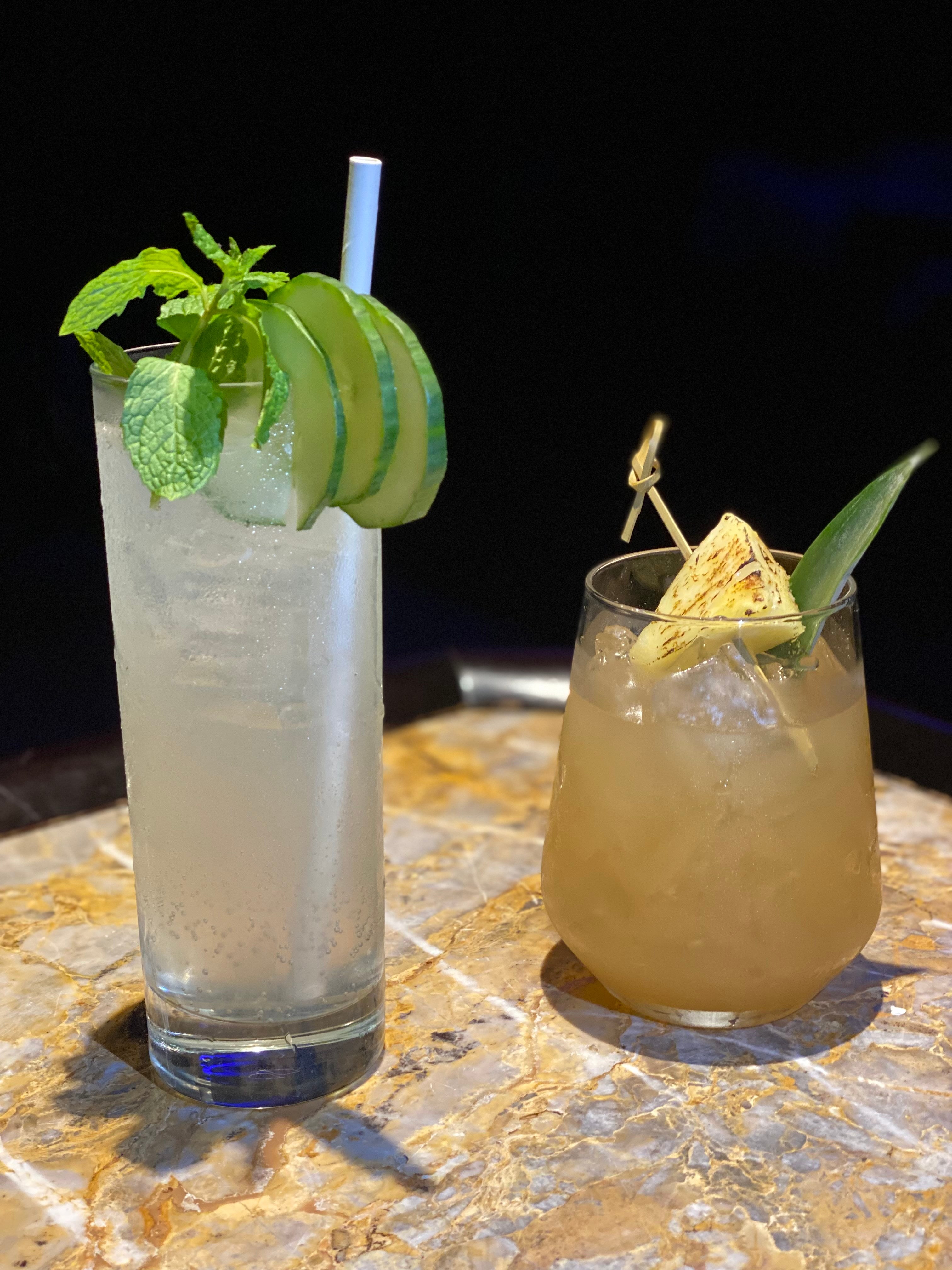 Cocktail in tall glass featuring mint and cucumber next to a short stemless cocktail with pineappe.
