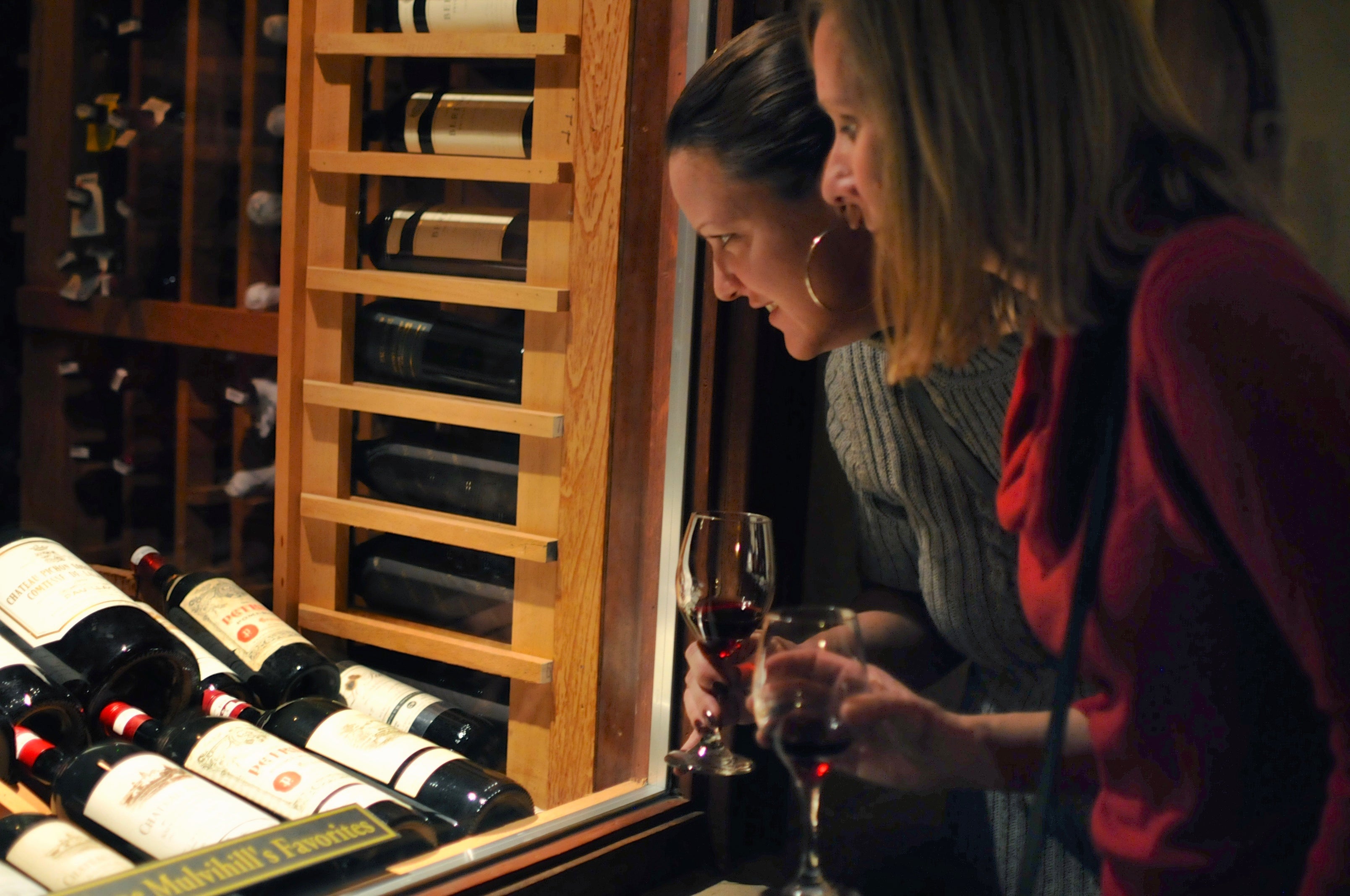 Two women look through glass window at wine collection.