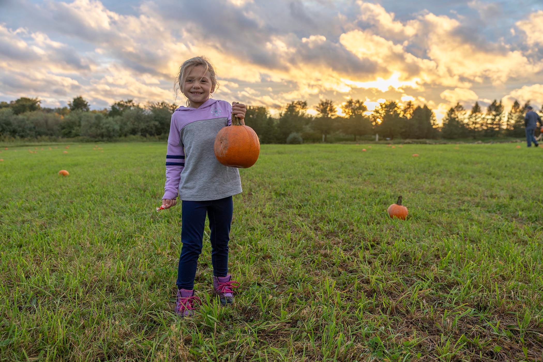 Young girl holding a pumpkin in an open field while the sun sets.