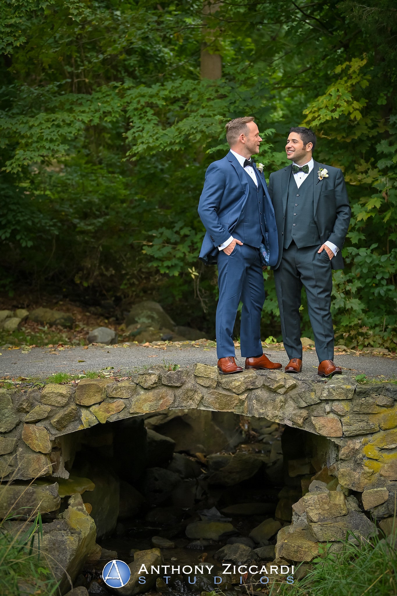 Grooms, wearing blue suits, looking at one another while standing in front of green foliage.