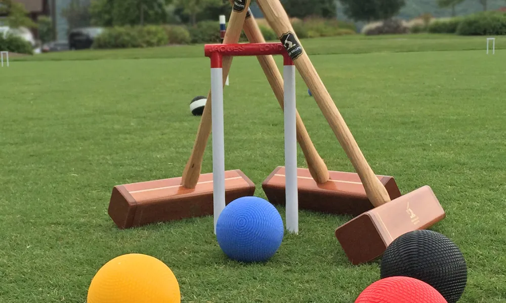 4 croquet balls on the grass and 3 croquet mallets leaning against a wicket