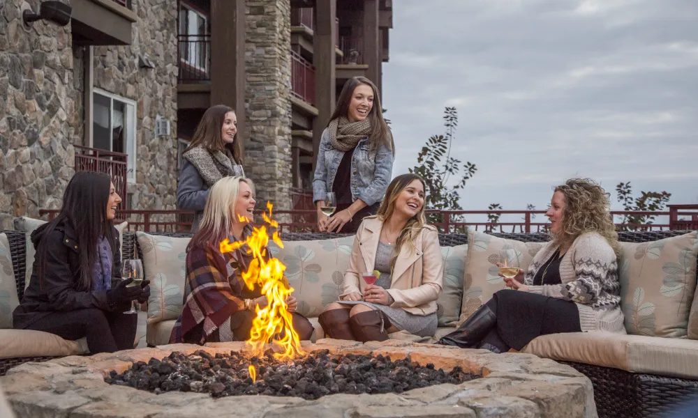 Group of 6 girlfriends laughing as they sit around firepit on fire and water terrace.