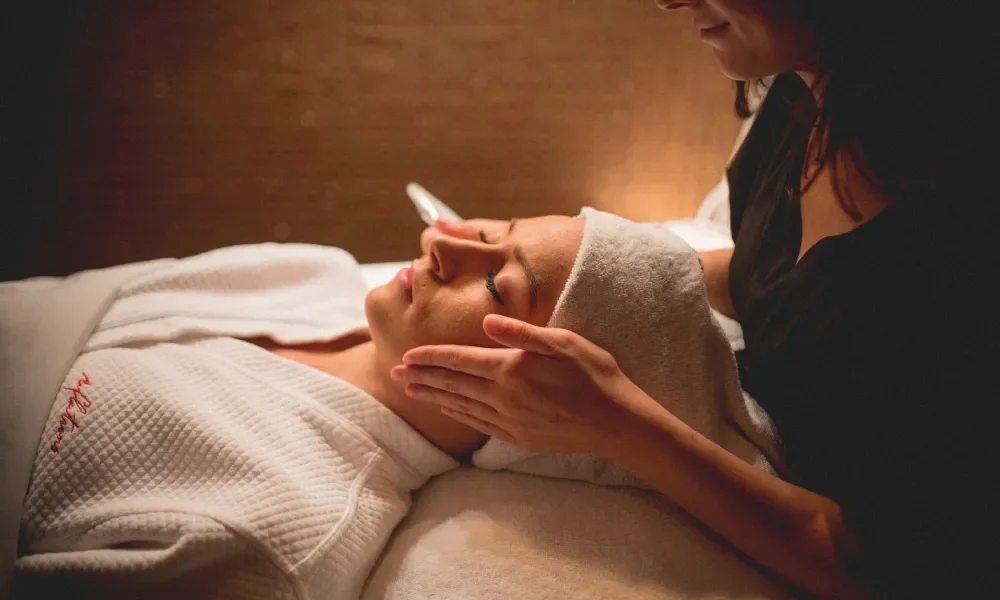 Woman in white robe and white hair towel laying on table with facialists hands on either side of her head.