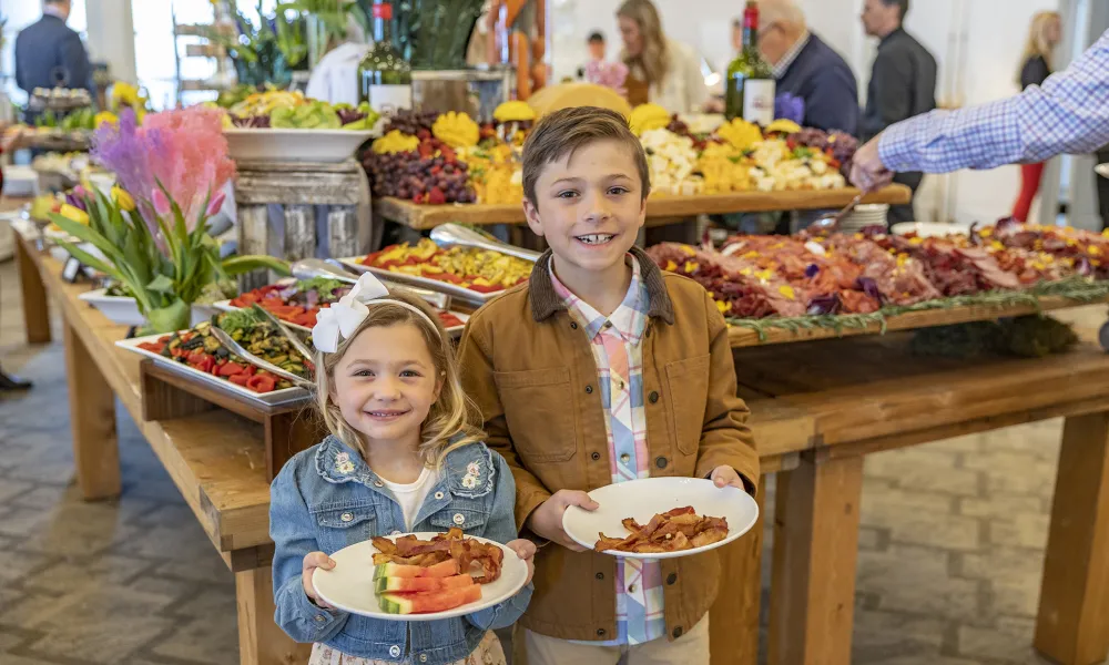 Two children holding plates of food in front of elaborate food display.