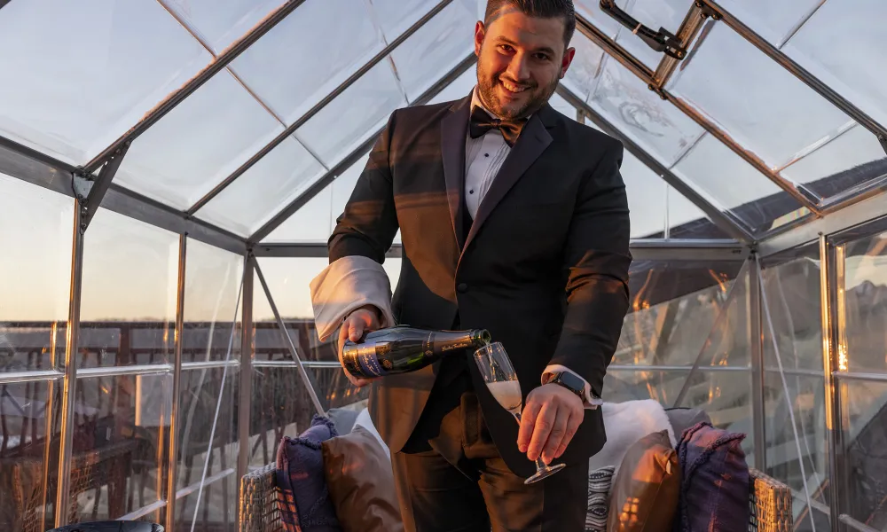 Waiter, dressed in a black suit,  pouring champagne while standing in the champagne chalet. 