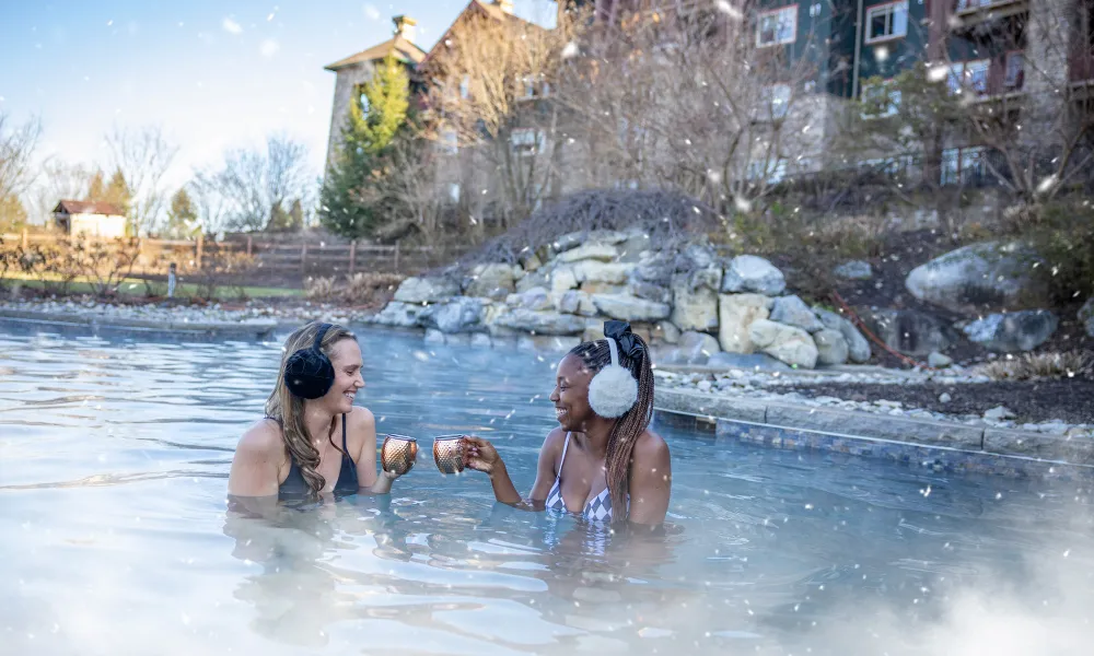Two women in outdoor snowpool wearing earmuffs and drinking mules.