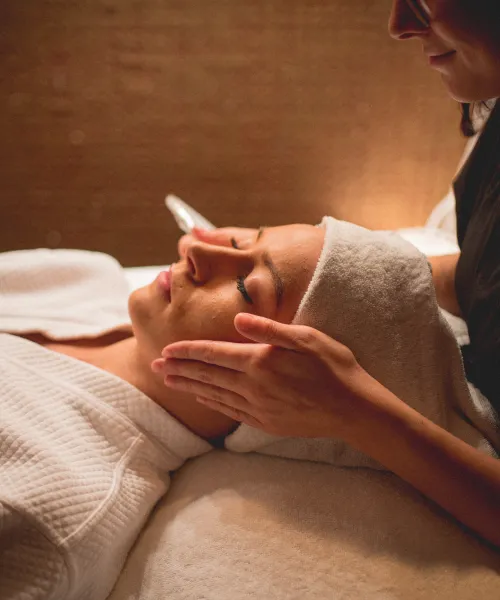 Woman in white robe and white hair towel laying on table with facialists hands on either side of her head.