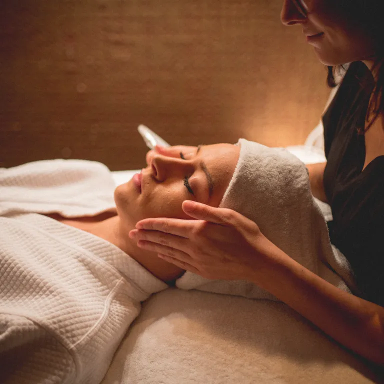 Woman in white robe and white hair towel laying on table with facialists hands on either side of her head.