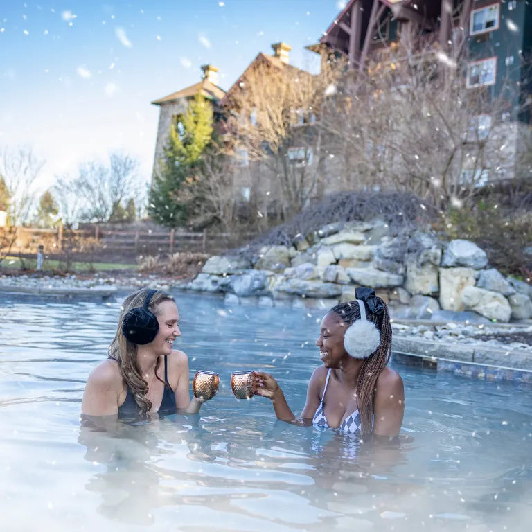 Two women in outdoor snowpool wearing earmuffs and drinking mules.