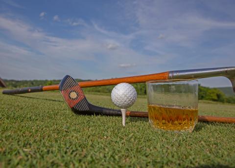 Old style golf clubs, golf ball on tee and whiskey glass on a golf course