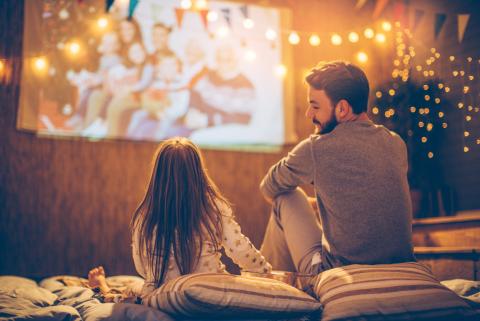 Father and daughter watching movie outdoors on screen under twinkly lights.